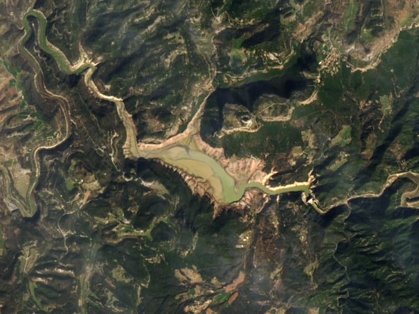 The same view of Sau Reservoir in March 2024, the water reduced to a narrow channel with pale exposed lakebed on either side.
